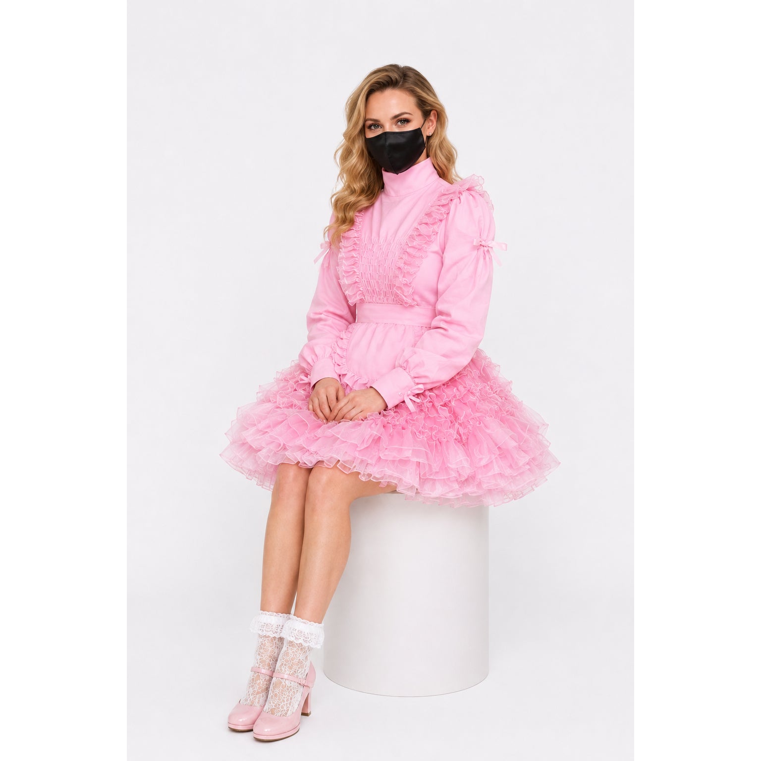 Pink sissy maid dress with ruffles, lace socks, and heels, worn by seated model on white stool