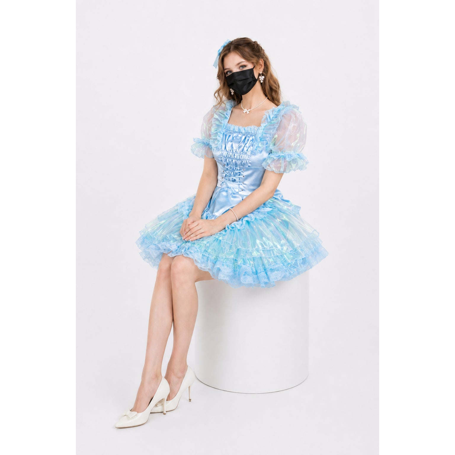 Woman in light blue sissy maid dress with ruffles, puff sleeves, and corset waist, seated on white pedestal