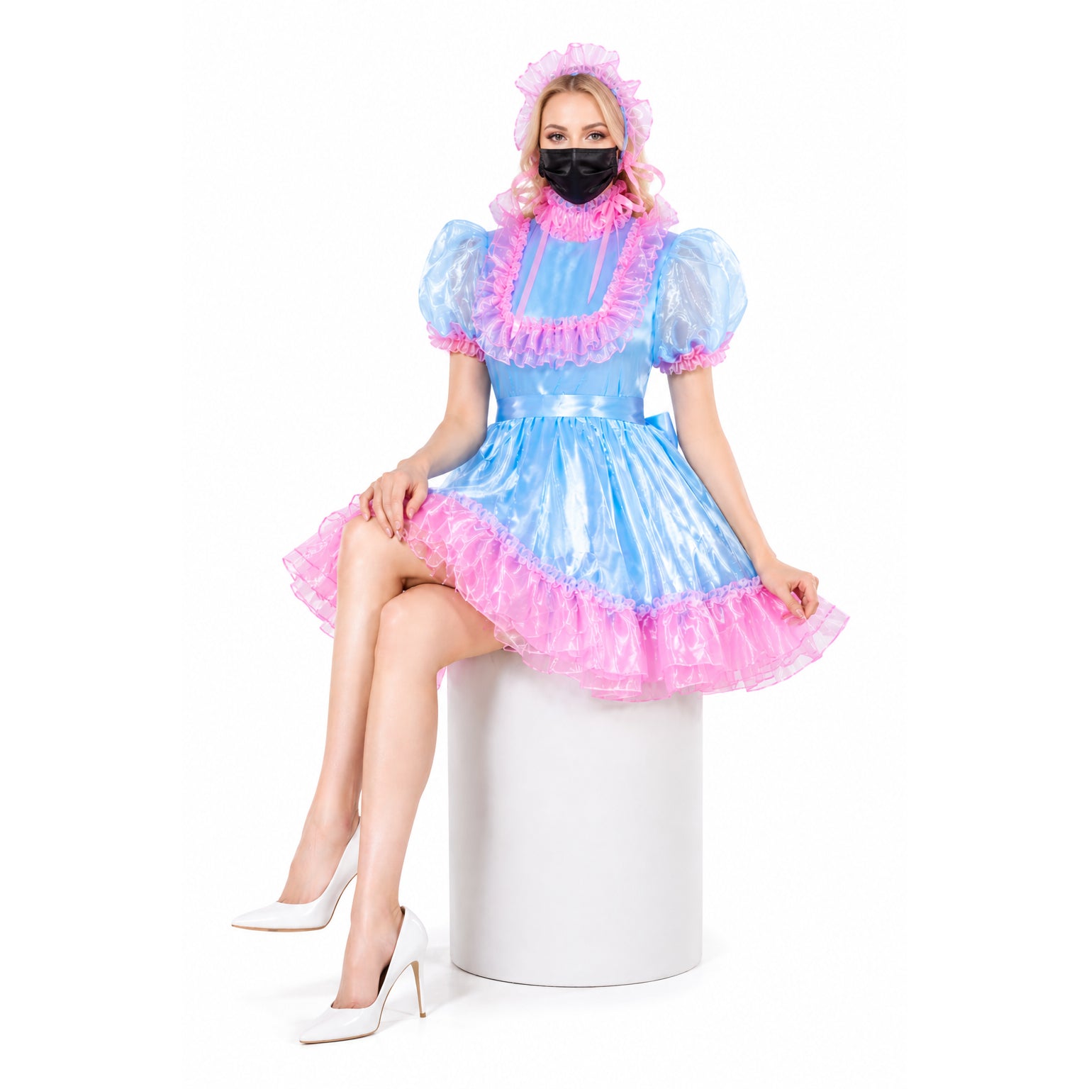 Blue and pink sissy maid dress with ruffles, worn with white heels and black mask, seated on white stool